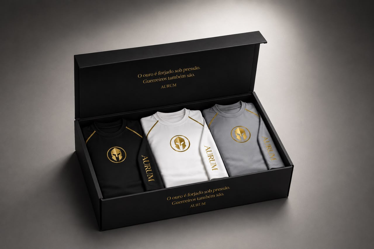 Caixa AURUM aberta com três rashguards — preta, branca e cinza — e a inscrição dourada: o ouro é forjado sob pressão, guerreiros também são